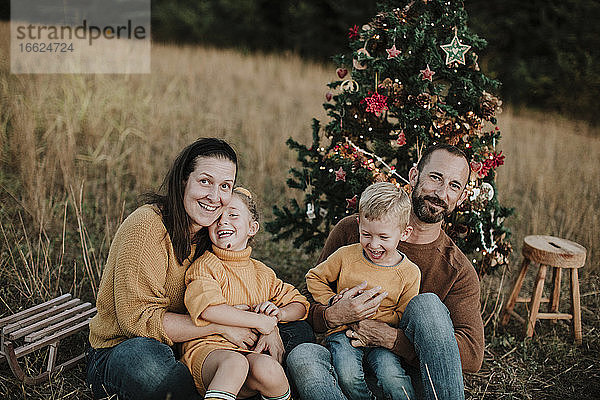 Fröhliche Familie mit Weihnachtsbaum bei Sonnenuntergang auf dem Land sitzend