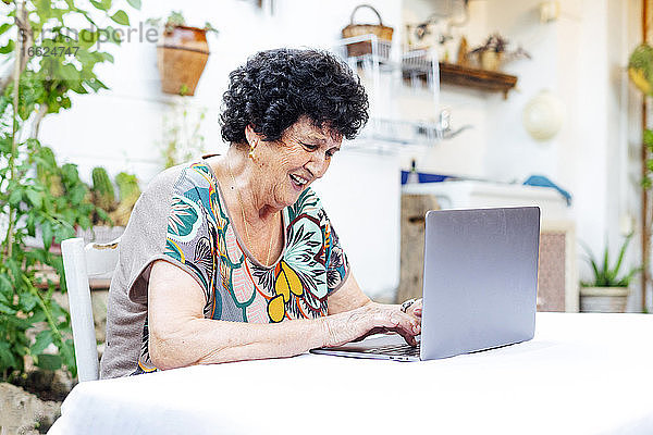 Glückliche ältere Frau  die einen Laptop auf dem Tisch benutzt  während sie im Hof sitzt
