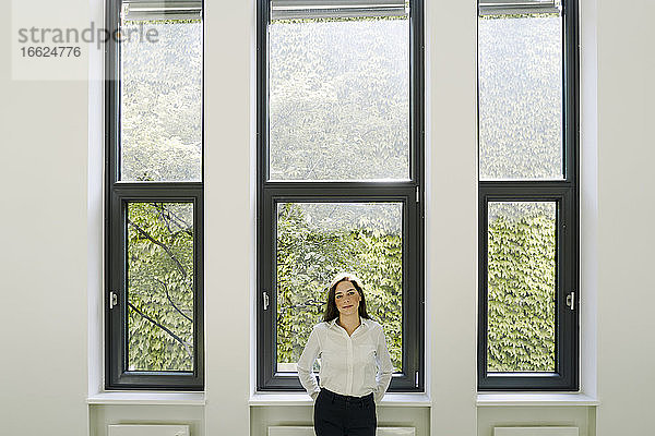 Geschäftsfrau mit Händen hinter dem Rücken steht gegen Fenster im Büro