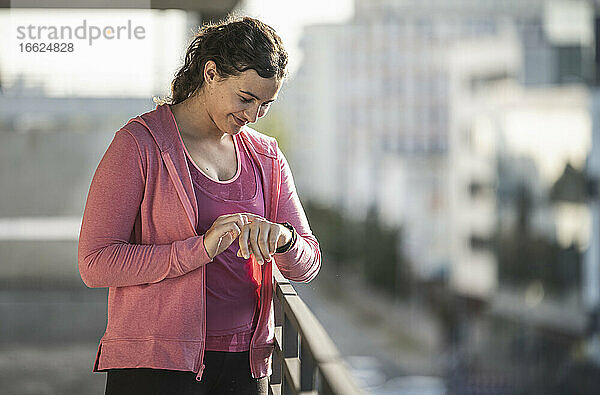 Lächelnde junge Frau prüft die Zeit  während sie an einem sonnigen Tag am Geländer steht