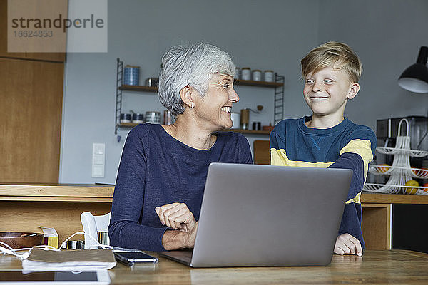 Lächelnder Enkel steht neben der Großmutter  die zu Hause am Laptop arbeitet