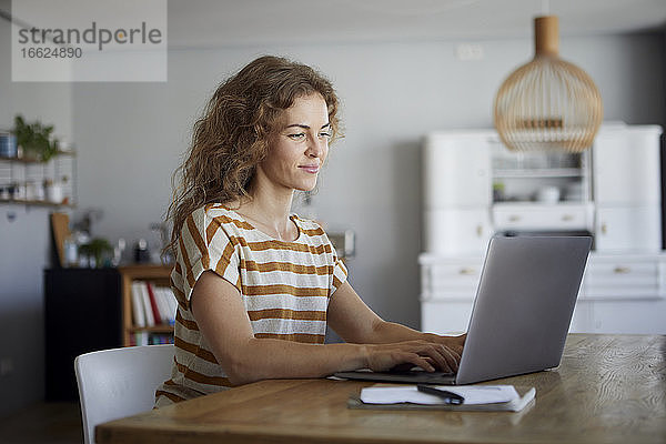 Mittlere erwachsene Frau arbeitet am Laptop  während sie zu Hause am Tisch sitzt