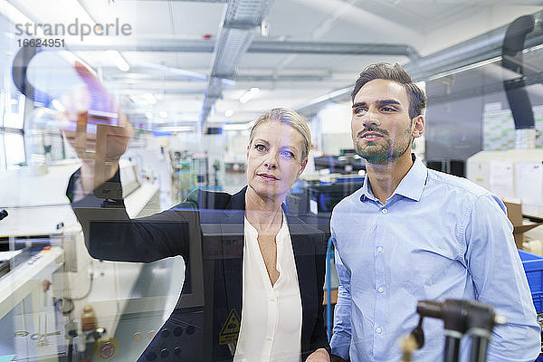 Selbstbewusste Geschäftsfrau  die mit einem jungen Ingenieur plant  während sie auf eine Glasoberfläche in einer Fabrik zeigt