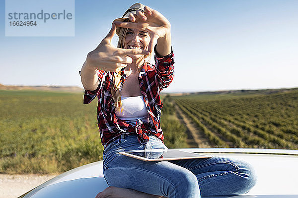 Lächelnde junge Frau zeigt Fingerrahmen  während sie auf dem Autodach sitzt