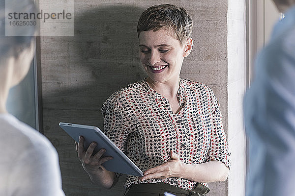Lächelnde Geschäftsfrau  die ein digitales Tablet hält  während sie sich mit Kollegen im Büro unterhält