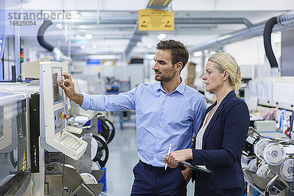 Reife Geschäftsfrau steht neben einem jungen männlichen Techniker  der in einer beleuchteten Fabrik Maschinen bedient