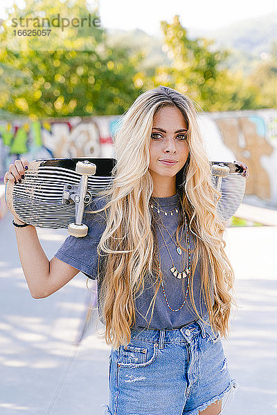 Selbstbewusste blonde Frau mit Skateboard im Park stehend