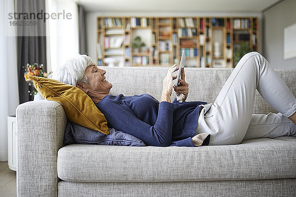 Ältere Frau schreibt Textnachrichten auf einem digitalen Tablet  während sie zu Hause auf dem Sofa liegt
