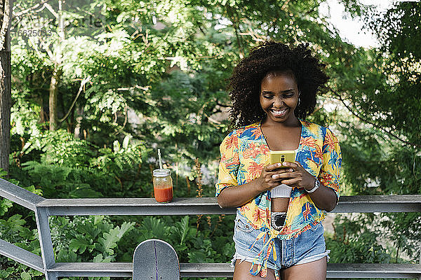 Lächelnde Frau  die mit ihrem Smartphone SMS schreibt  steht an einem Geländer im Park
