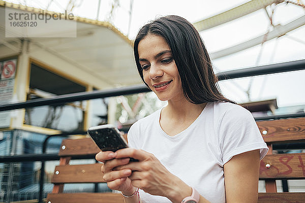 Nahaufnahme einer lächelnden Frau  die ein Mobiltelefon benutzt  während sie auf einer Bank im Vergnügungspark sitzt