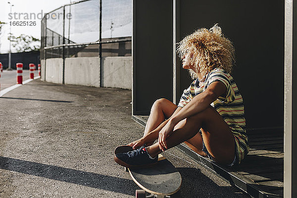 Nachdenkliche Frau mit Skateboard sitzend und wegschauend an einem sonnigen Tag