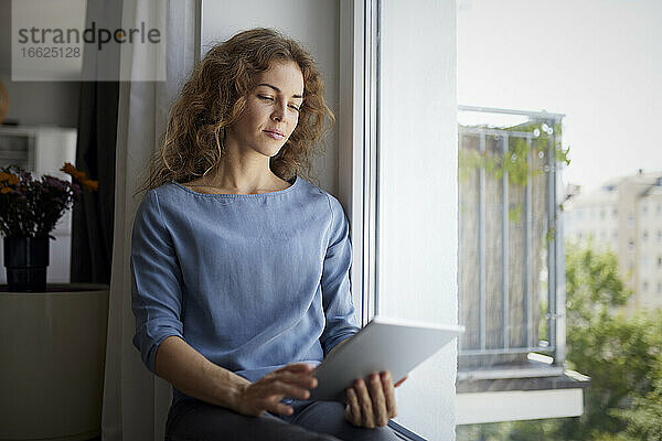 Frau benutzt digitales Tablet  während sie zu Hause auf der Fensterbank sitzt