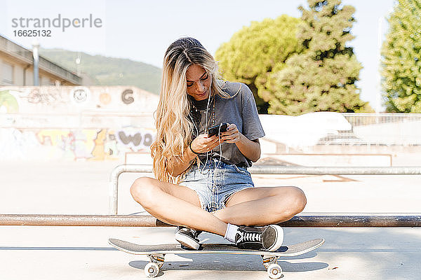 Blonde Frau  die ihr Smartphone benutzt  während sie mit dem Skateboard im Park an einem sonnigen Tag sitzt
