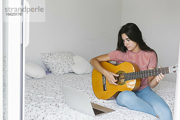 Junge Frau lernt online Gitarre  während sie zu Hause im Schlafzimmer sitzt