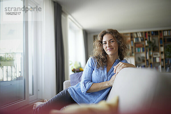 Lächelnde Frau  die zu Hause auf dem Sofa am Fenster sitzt