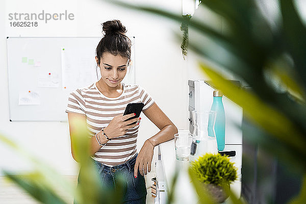 Junge weibliche Designerin  die mit ihrem Smartphone Textnachrichten verschickt  während sie an einem Tisch im Kreativbüro steht