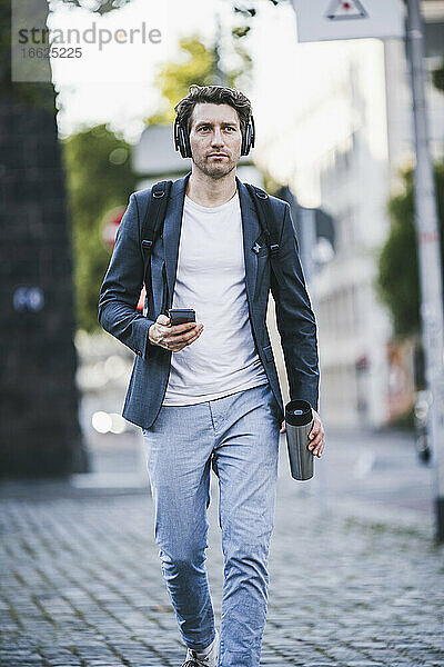Geschäftsmann mit Kopfhörer  der ein Mobiltelefon benutzt  während er auf einer Straße in der Stadt geht