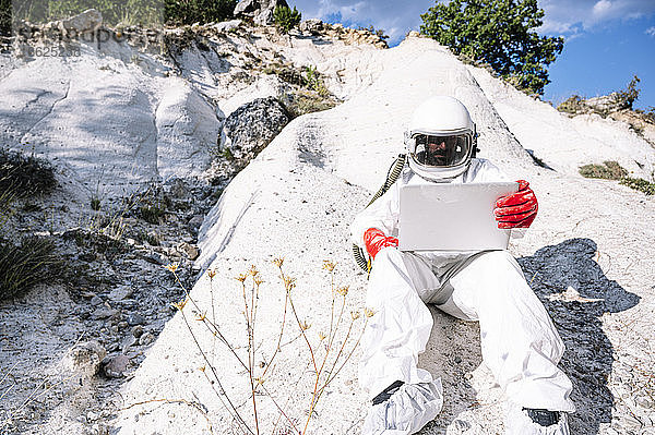 Männlicher Astronaut  der einen Laptop benutzt  während er auf einem Berg sitzt