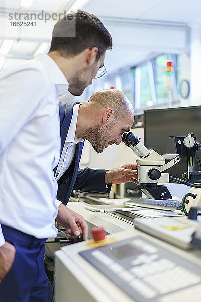 Junger Techniker  der neben einem männlichen Kollegen steht und durch ein Mikroskop im Labor schaut