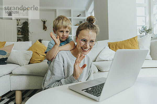 Mutter und Sohn winken mit der Hand zum Videoanruf auf dem Laptop  während sie zu Hause sitzen