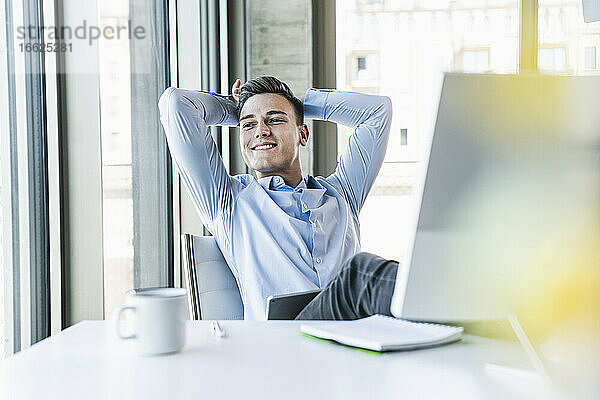 Nachdenklicher männlicher Fachmann mit Händen hinter dem Kopf am Schreibtisch sitzend im Büro