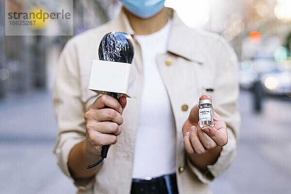 Nahaufnahme einer Reporterin mit Maske  die eine Impfstoffampulle zeigt  während sie im Freien steht