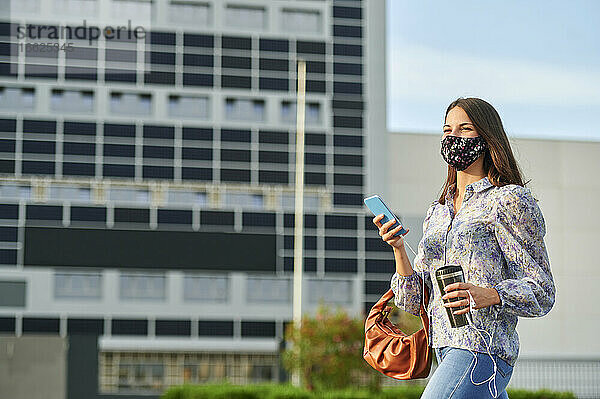 Junge Frau mit Gesichtsmaske  die beim Spaziergang in der Stadt ein Mobiltelefon benutzt