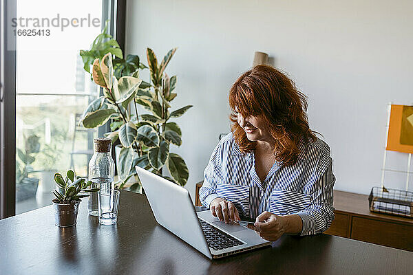 Reife Frau sitzt am Tisch und arbeitet zu Hause am Laptop