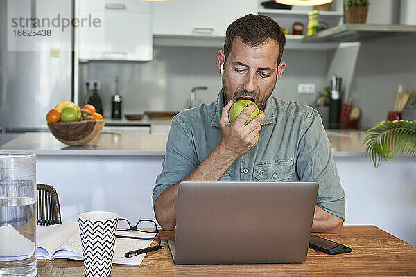 Gut aussehender männlicher Freiberufler  der einen frischen Apfel isst  während er zu Hause mit seinem Laptop arbeitet