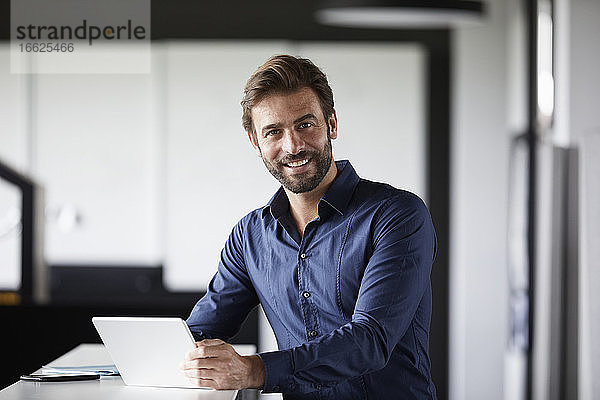 Lächelnder Geschäftsmann  der ein digitales Tablet benutzt  während er am Schreibtisch im Büro steht
