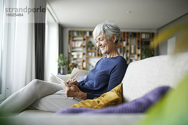 Lächelnde Frau  die in ein digitales Tablet schaut  während sie zu Hause auf dem Sofa sitzt
