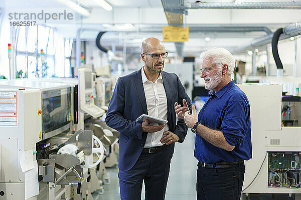 Ein Geschäftsmann hält ein digitales Tablet in der Hand und schaut auf einen leitenden Manager  der in einer beleuchteten Fabrik diskutiert