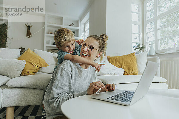 Junge zeigt auf Laptop  während er zu Hause hinter seiner Mutter steht