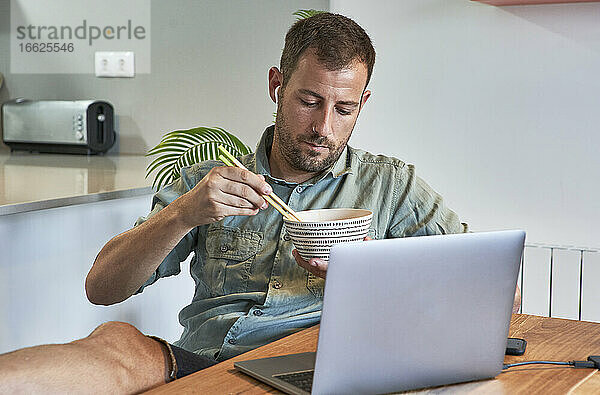 Entspannter Freiberufler isst Nudeln  während er mit seinem Laptop am Esstisch sitzt