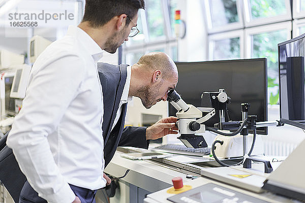 Junger männlicher Techniker steht neben einem Geschäftsmann  der durch ein Mikroskop im Labor schaut