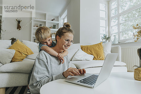 Frau arbeitet am Laptop  während ein Junge sie zu Hause von hinten umarmt