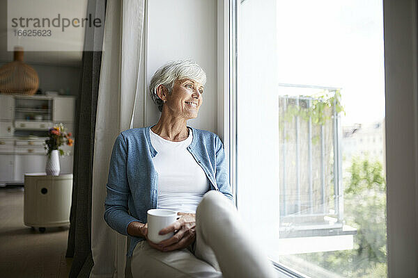 Ältere Frau schaut aus dem Fenster  während sie zu Hause sitzt