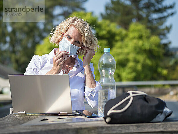 Frau mit Maske sitzt mit Laptop im Hinterhof