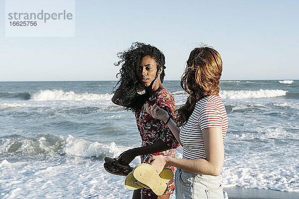 Freunde halten Pantoffel  während sie am Strand vor dem Wasser stehen