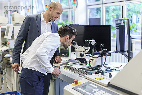 Junger Ingenieur schaut durch ein Mikroskop  während er neben einem Geschäftsmann im Labor steht