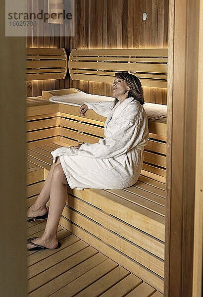 Nachdenkliche Frau im Bademantel auf einer Holzsauna im Wellnessbereich sitzend