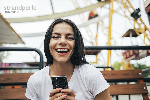 Frau hält Smartphone lachend auf einer Bank im Vergnügungspark sitzend