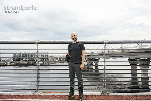Lächelnder junger Mann mit Inline-Skates  der auf einer Brücke stehend sein Smartphone benutzt
