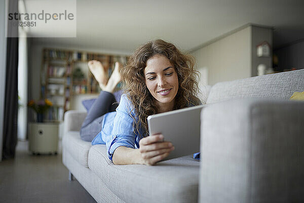 Frau benutzt digitales Tablet  während sie zu Hause auf dem Sofa liegt
