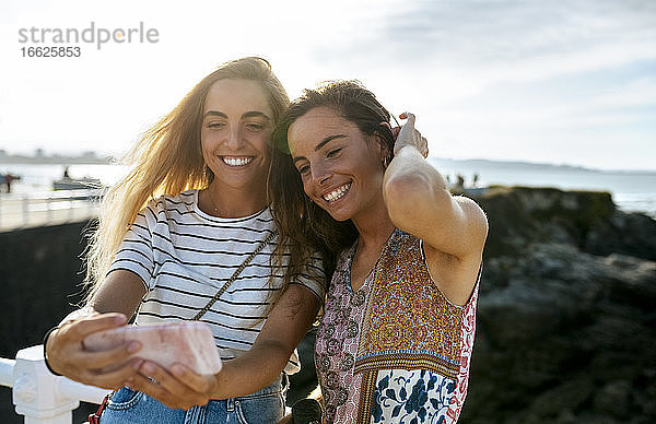 Lächelnde junge Schwestern machen am Wochenende ein Selfie am Strand