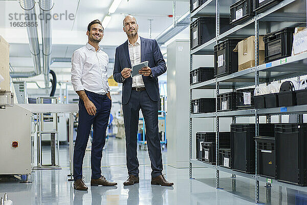 Geschäftsmann hält ein digitales Tablet in der Hand  während er neben einem lächelnden Ingenieur steht und auf eine Fabrik blickt