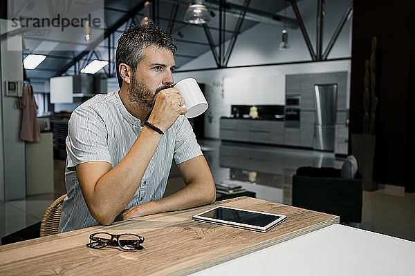 Nachdenklicher männlicher Unternehmer trinkt Kaffee und sitzt mit digitalem Tablet und Brille am Schreibtisch im Büro