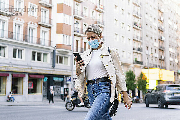 Junge Frau mit Maske  die ein Mobiltelefon benutzt  während sie auf einer Straße in der Stadt spazieren geht