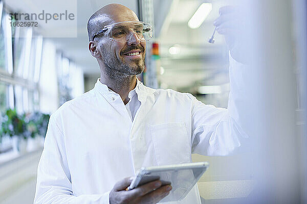 Lächelnder männlicher Wissenschaftler  der eine Probe betrachtet und ein digitales Tablet in einer Fabrik hält