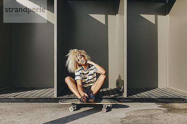 Frau mit Skateboard  die an einem sonnigen Tag gegen Metallhütten sitzt